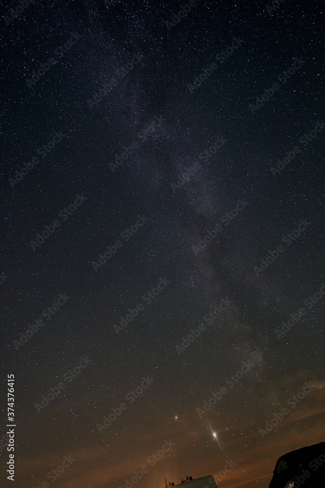 Milky Way in front of the blue night sky, the planet Jupiter can be seen, vehicel parts of two small transporters are in the foreground, the horizon shines orange in the lower part. Germany, Heroldsta