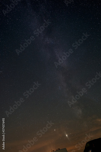 Milky Way in front of the blue night sky, the planet Jupiter can be seen, vehicel parts of two small transporters are in the foreground, the horizon shines orange in the lower part. Germany, Heroldsta