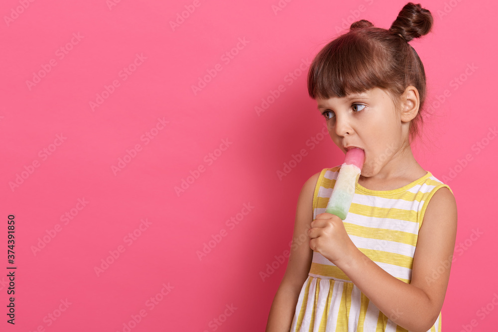 Adorable little girl with knots in yellow and white summer dress eating ice cream on pink background, copy space for advertisement or promotional text. Adorable little girl with knots in yellow and white summer dress eating ice cream on pink background, copy space for advertisement or promotional text.