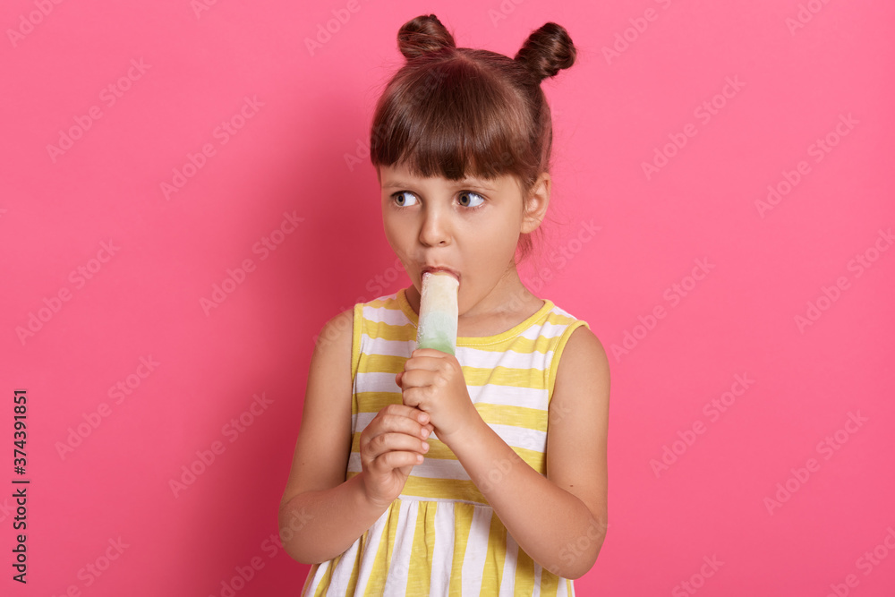 Charming little girl with knots wearing dress, eating ice cream, posing isolated over pink background, looking aside, dresses summer outfit, having knots. Charming little girl with knots wearing dress, eating ice cream, posing isolated over pink background, looking aside, dresses summer outfit, having knots.