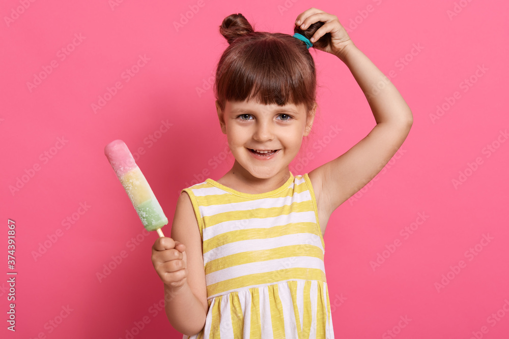 Small little child with charming smile, hair bun, dressed yellow and white striped sarafan, looks at camera, poses isolated over pink background, touching her knot. Small little child with charming smile, hair bun, dressed yellow and white striped sarafan, looks at camera, poses isolated over pink background, touching her knot.