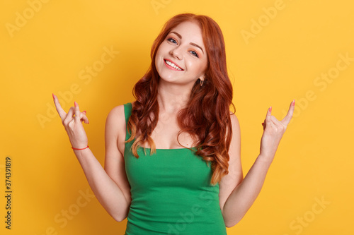 Emotional female makes rock n roll gesture, enjoys cool music at party, demonstrates hand gesture, dressed green outfit, models over yellow background.