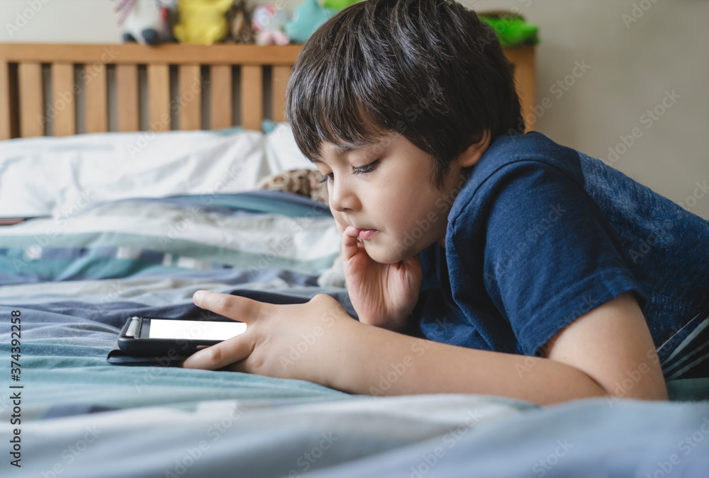 Kid using tablet for his homework, Child lying in bed playing game on digital touch pad in bed room,Young boy lying down with mock up tablet.Home schooling, Social Distance,E-learning online education Kid using tablet for his homework, Child lying in bed playing game on digital touch pad in bed room,Young boy lying down with mock up tablet.Home schooling, Social Distance,E-learning online education