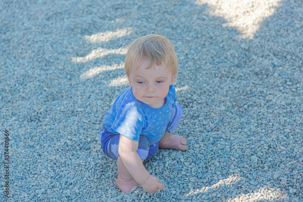 Fototapeta premium Funny baby sitting on the ground, covered with small stones. A blond boy plays with pebbles in a city Park.