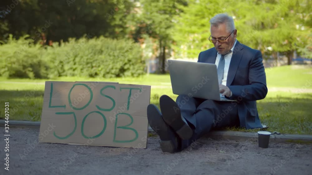 Businessman sitting on street with lost job cardboard sign and paper cup for charity and sing laptop Businessman sitting on street with lost job cardboard sign and paper cup for charity and sing laptop