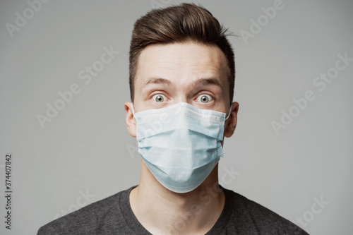 portrait of a young man of Caucasian appearance in a blue mask looks at the camera. grey t-shirt, grey background. a protective agent against the pandemic face. emotion surprise