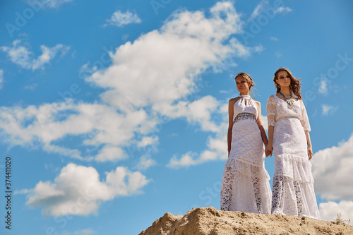 Two young girls in white Boho dresses pose together on a sand hill against a blue sky. Happy young people, teenage friendship