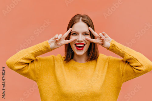 Blithesome young woman in yellow clothes posing with peace sign. Indoor photo of good-humoured brown-haired lady smiling in studio.