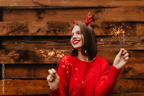 Pleasant girl in winter red sweater holding bengal light. Outdoor shot of happy european lady with short hair posing with sparklers.