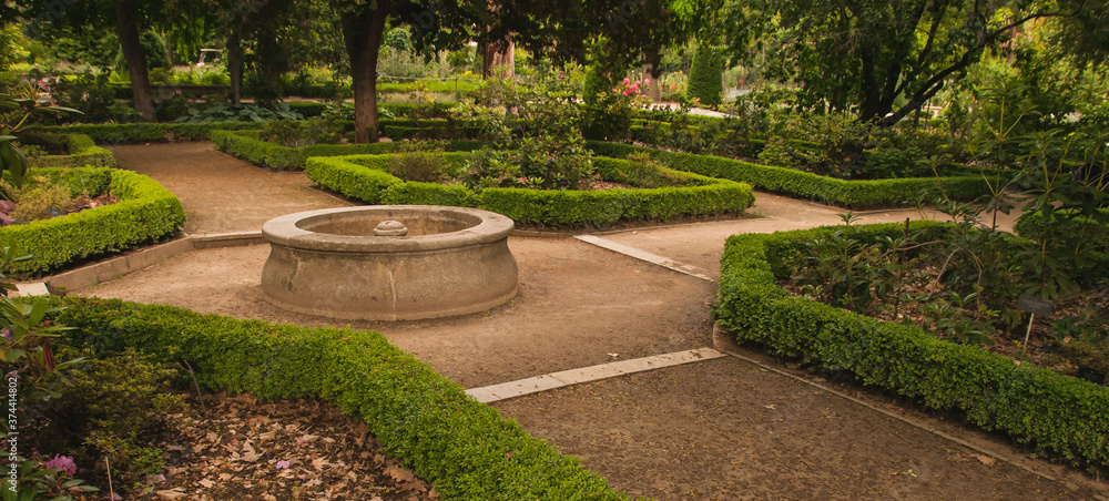 Obraz premium Fuente redonda de piedra situada en un cruce de caminos dentro de un jardín botánico, rodeada de parterres con diferentes tipos de plantas y árboles.