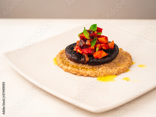 Ratatouille Stuffed Portobello Mushroom on Top of Creamy Quinotto with Olive Oil Sprinkles and Peppercorn served on Square White Plate photo