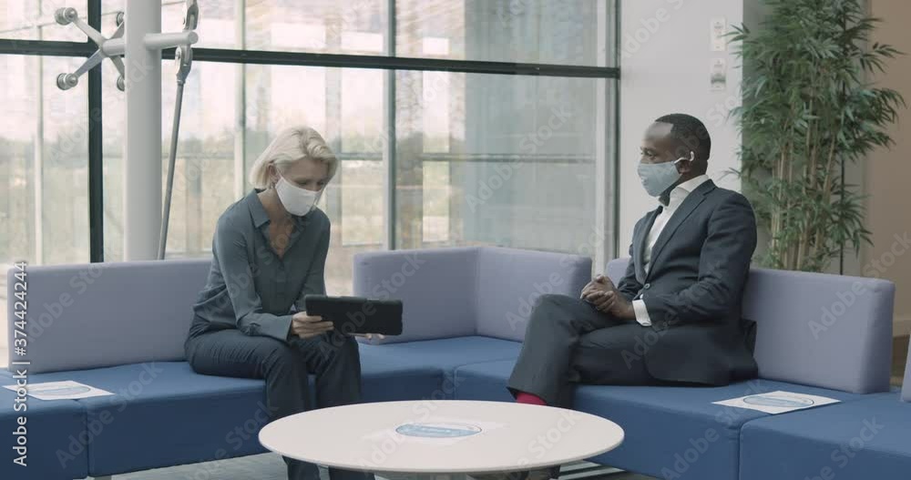 Business people in office meeting on sofa wearing face mask with digital tablet in lobby Business people in office meeting on sofa wearing face mask with digital tablet in lobby
