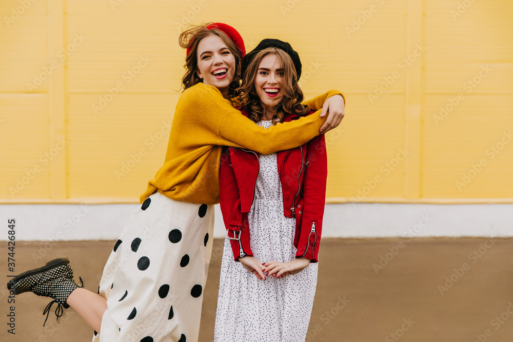 Long-haired brunette woman in beret enjoying photoshoot with friend in autumn day. Magnificent ladies expressing love on yellow background.
