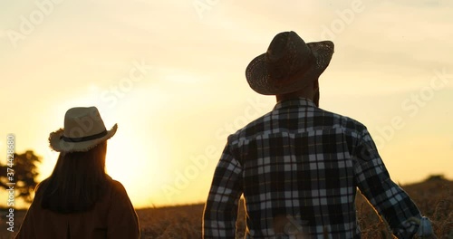Close up portrait of Caucasian couple of man and woman walking in golden wheat field on sunset. Rear of pretty girl and handsome male in hats outdoors on farm. Lovely couple concept