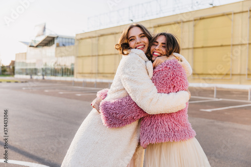 Graceful brown-haired lady embracing friend during autumn photoshoot on the street. Portrait of good-humoured girls spending weekend together.