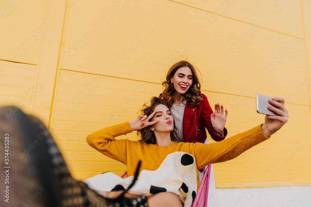 Fototapeta premium Charming girl in red jacket posing with friend. Lovely ladies making selfie outdoor.