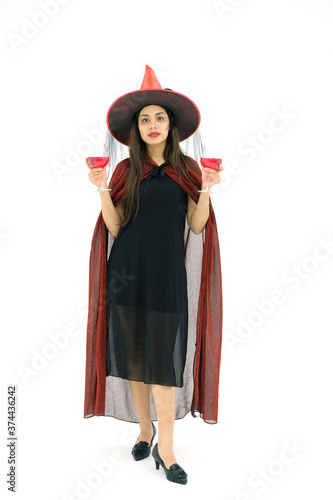 A young woman with a halloween party costume stands on a white background holding a red wide glass.