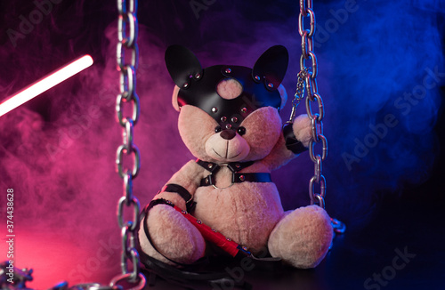 toy bear dressed in leather belts harness accessory for BDSM games on a dark background in neon light