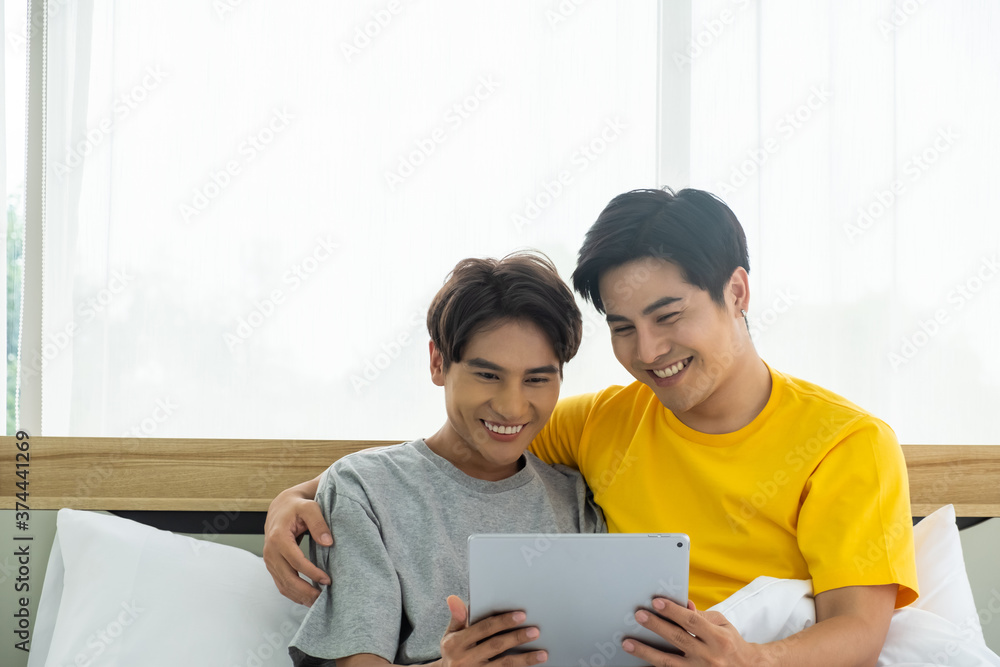 Selective focus at face. Good looking Asian gay homosexual couple hug and smile while using Digital tablet smartphone, showing love expression on the bed. LGBT relationship concept with copy space. Selective focus at face. Good looking Asian gay homosexual couple hug and smile while using Digital tablet smartphone, showing love expression on the bed. LGBT relationship concept with copy space.