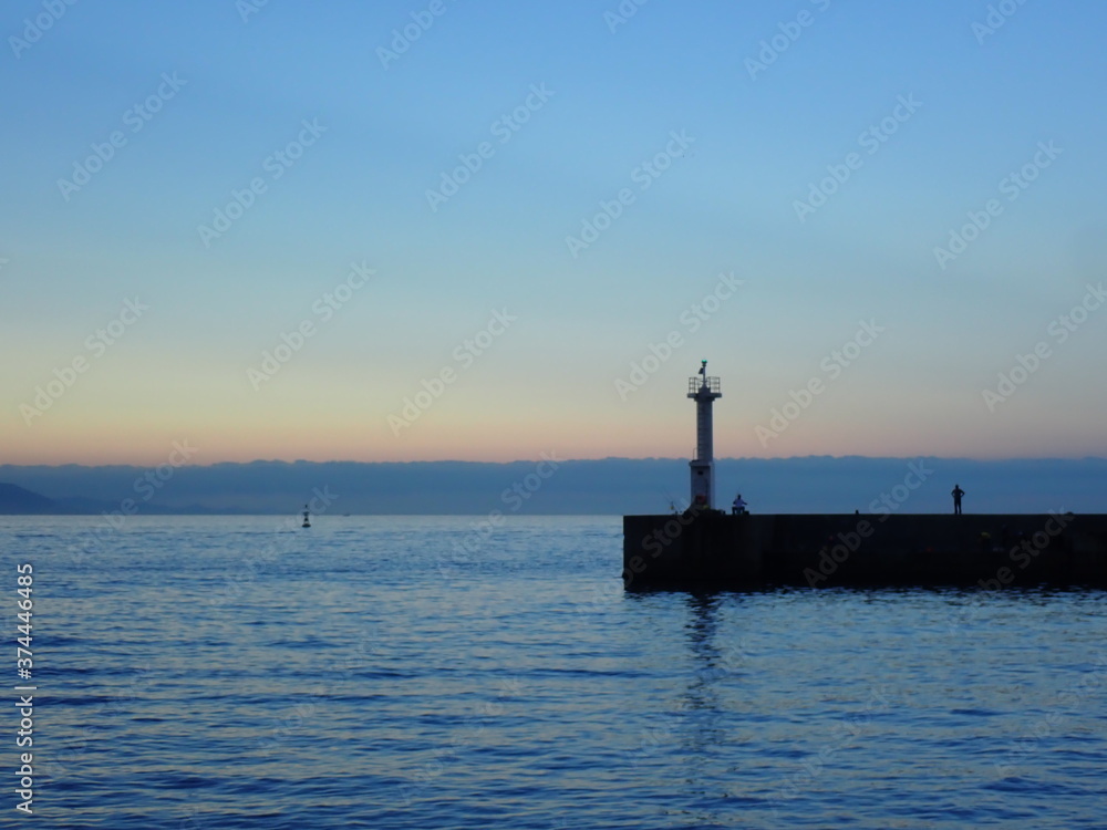 夜明けの空と灯台のもとで釣りをする人と海を眺める人