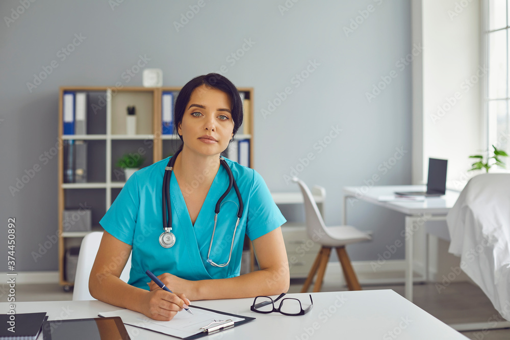 Woman doctor therapist sitting in medical clinic office, making notes and looking at camera Woman doctor therapist sitting in medical clinic office, making notes and looking at camera