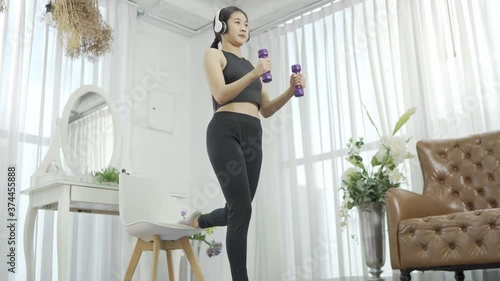 An Asian woman wearing a black sports bar and white headphones. Squatting with dumbbells on the balcony in the living room of the house.
 photo