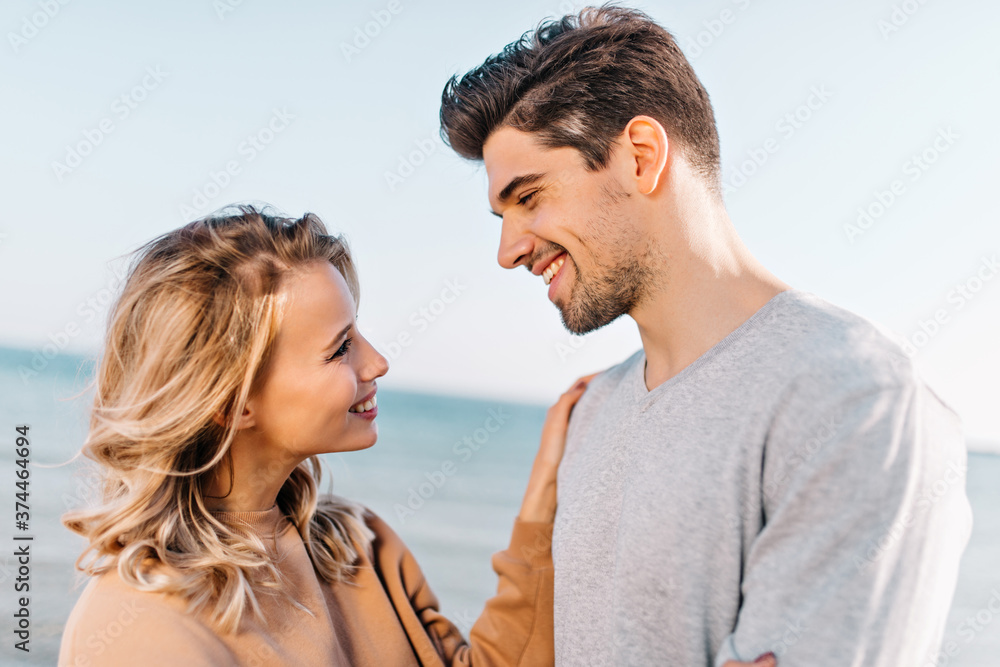 Interested curly caucasian girl looking at boyfriend with love. Smiling couple posing at sea photoshoot. Interested curly caucasian girl looking at boyfriend with love. Smiling couple posing at sea photoshoot.