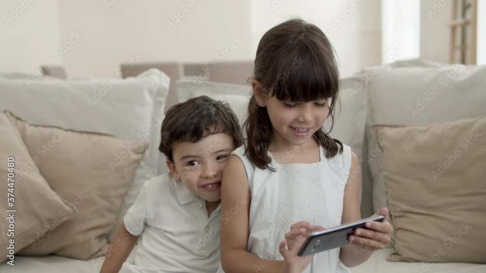 Sweet little kids using phone for video talk, sister dialing number and waving hello at webcam, while brother getting naughty. Medium shot. Communication concept Sweet little kids using phone for video talk, sister dialing number and waving hello at webcam, while brother getting naughty. Medium shot. Communication concept