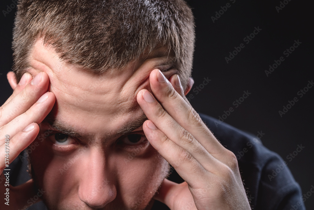 Fototapeta premium Young handsome man in a black shirt sits and holds his head on a black background. Experiencing stress, pain