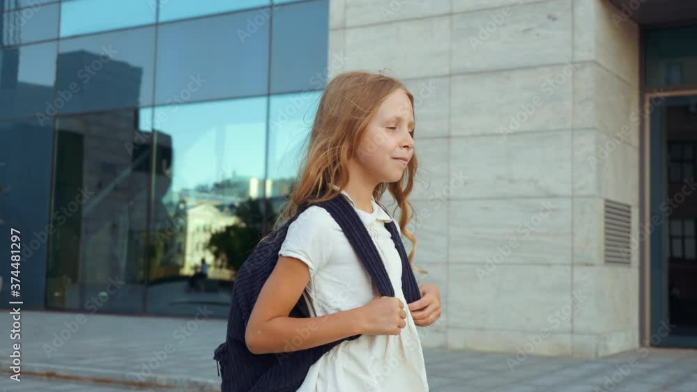 Cute little schoolgirl with golden hair carries large backpack, walks home from school through city, jumps happily, runs along road. Pupil elementary grades goes to class lesson to classmates, smiling Cute little schoolgirl with golden hair carries large backpack, walks home from school through city, jumps happily, runs along road. Pupil elementary grades goes to class lesson to classmates, smiling