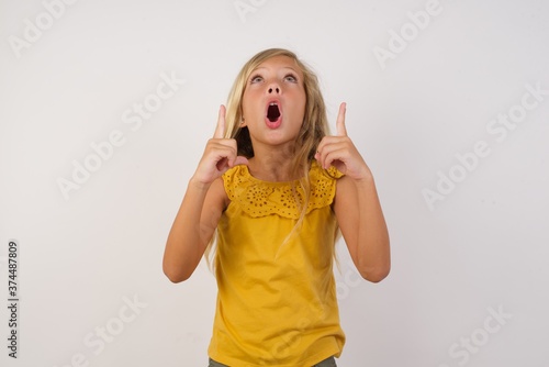 Young blonde kid girl wearing yellow dress over white background amazed and surprised looking up and pointing with fingers and raised arms.