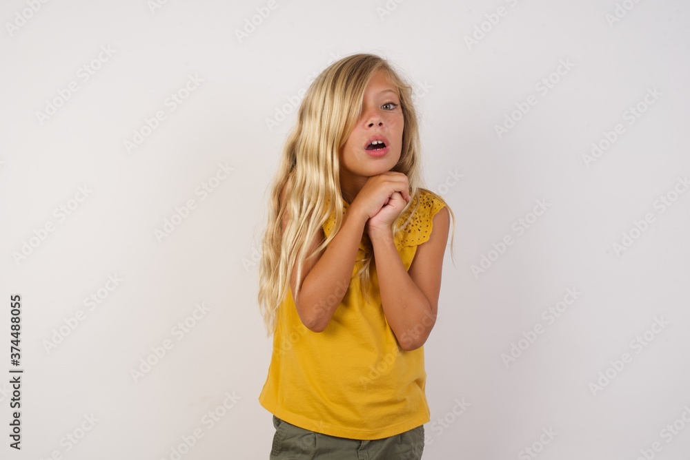 Little girl with beautiful blonde hair over white background praying for luck has hands crossed near face, amazed and opening mouth looking front. Little girl with beautiful blonde hair over white background praying for luck has hands crossed near face, amazed and opening mouth looking front.