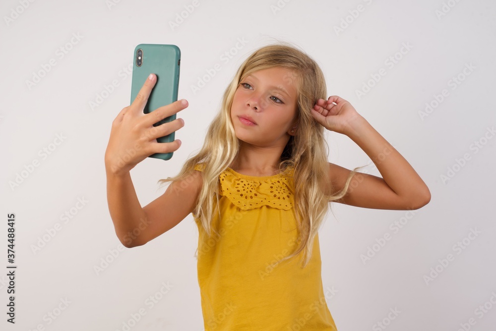 Photo of charming Little girl with beautiful blonde hair over white background smiling and taking a selfie ready to post it on her social media. Photo of charming Little girl with beautiful blonde hair over white background smiling and taking a selfie ready to post it on her social media.