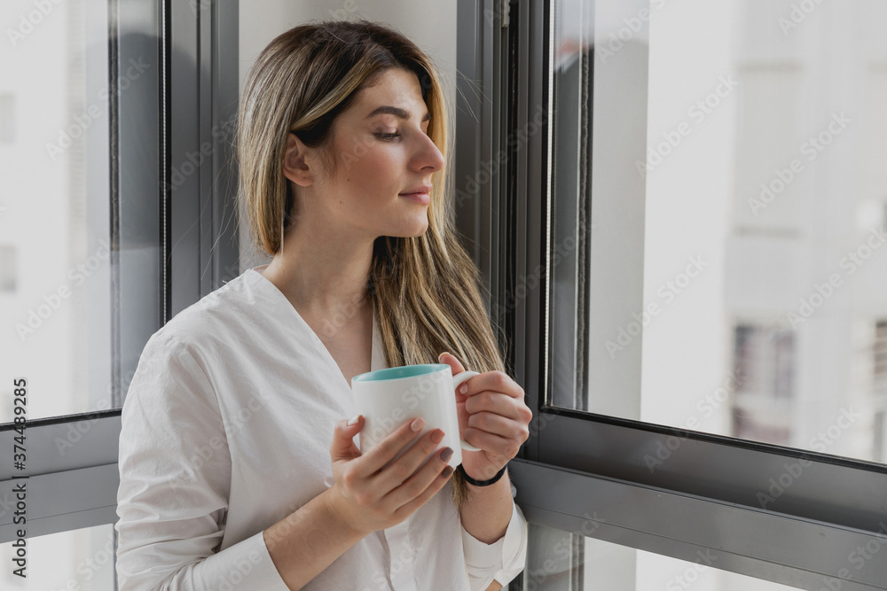 Young beautiful woman near window with cup of coffee or tea at home. Dream and relax. Good day. Woman in the morning is holding a mug and looking at the sunrise and having a perfect cozy morning Young beautiful woman near window with cup of coffee or tea at home. Dream and relax. Good day. Woman in the morning is holding a mug and looking at the sunrise and having a perfect cozy morning