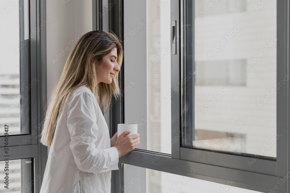 Young beautiful woman near window with cup of coffee or tea at home. Dream and relax. Good day. Woman in the morning is holding a mug and looking at her child in the playground from home Young beautiful woman near window with cup of coffee or tea at home. Dream and relax. Good day. Woman in the morning is holding a mug and looking at her child in the playground from home