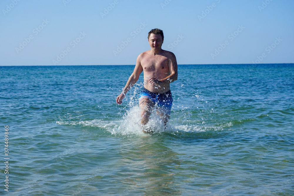 The guy runs along the waves in the sea with azure water. Splashes fly to the sides