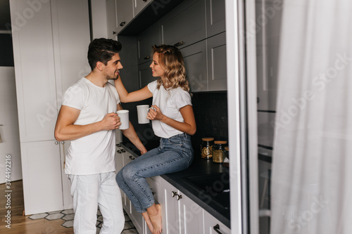 Pleasant girl touching husband's face. Indoor portrait of caucasian couple drinking coffee in kitchen.