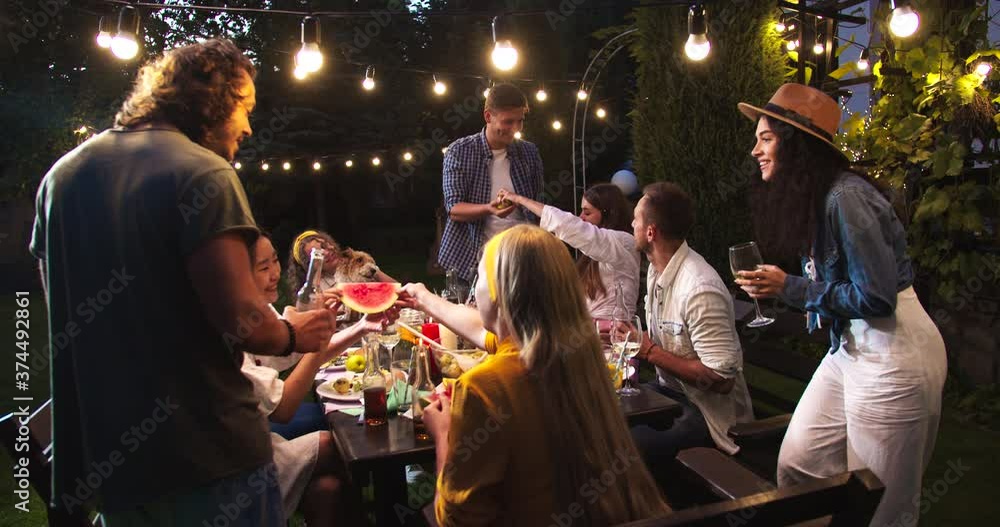 Happy multi-ethnic people having fun at garden at night. Caucasian boys and girls drinking and eating while talking outdoor. African American female laughing. Asian girl with watermelon. Party concept Happy multi-ethnic people having fun at garden at night. Caucasian boys and girls drinking and eating while talking outdoor. African American female laughing. Asian girl with watermelon. Party concept