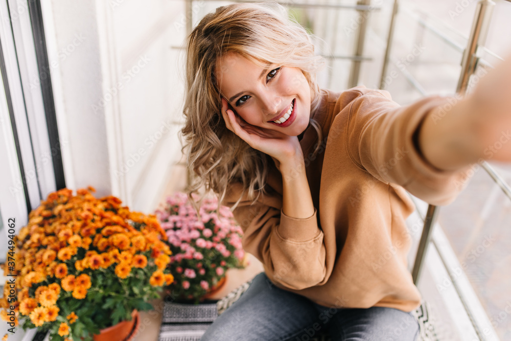 Carefree young woman making selfie near yellow flowers. Cute caucasian girl sitting on balcony. Carefree young woman making selfie near yellow flowers. Cute caucasian girl sitting on balcony.