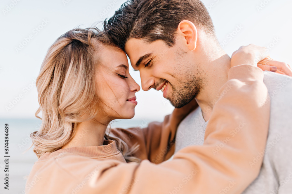 Fototapeta premium Elegant girl embracing husband with eyes closed. Outdoor photo of romantic couple posing on the street.