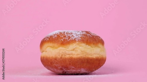 Nice round donut with filling. Powdered sugar flies from above. On a pink background. Pastry product, contains a lot of sugar.