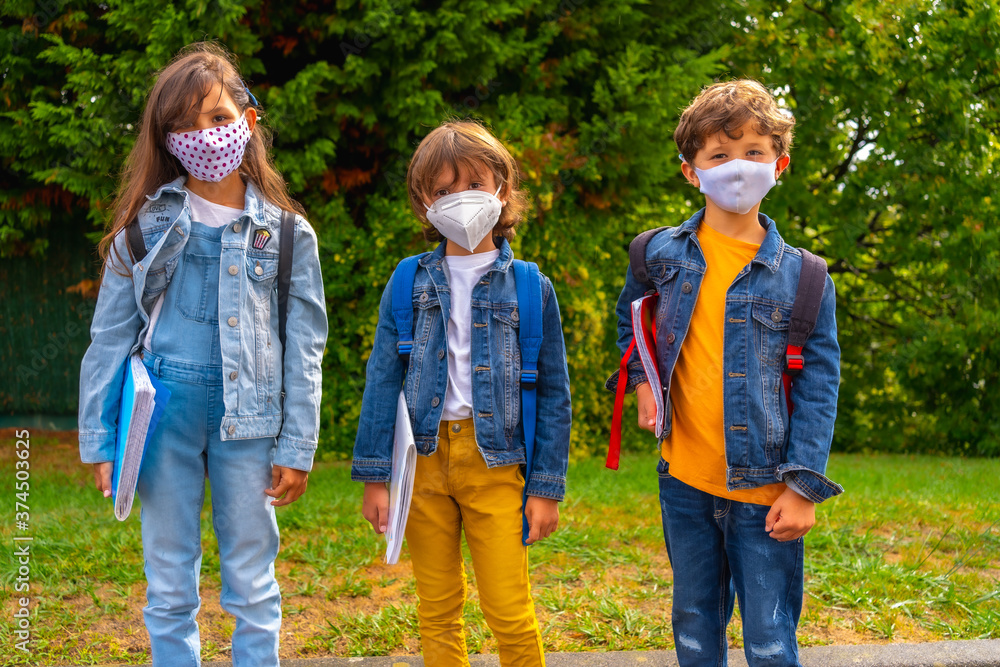 Three brothers with face masks ready to go back to school. New normality, social distance, coronavirus pandemic, covid-19. Waiting to go to school with green plants in the background Three brothers with face masks ready to go back to school. New normality, social distance, coronavirus pandemic, covid-19. Waiting to go to school with green plants in the background