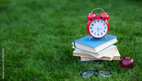 Red Alarm clock and stack old books in park in autumn morning, concept of education and reading. Books, apple, glasses with an alarmclock lie on grass near school. copy space. mockup. Back to school.	