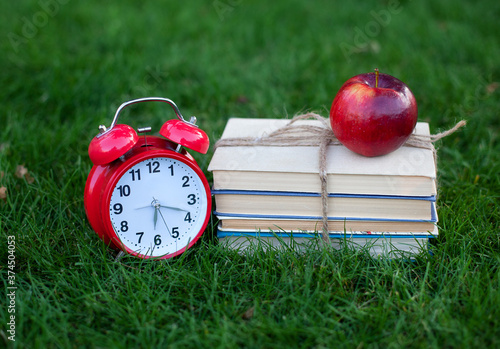 Red Alarm clock and stack old books in park in autumn morning, concept of education and reading. Books, apple with an alarmclock lie on grass near school. copy space. mockup. Back to school. 