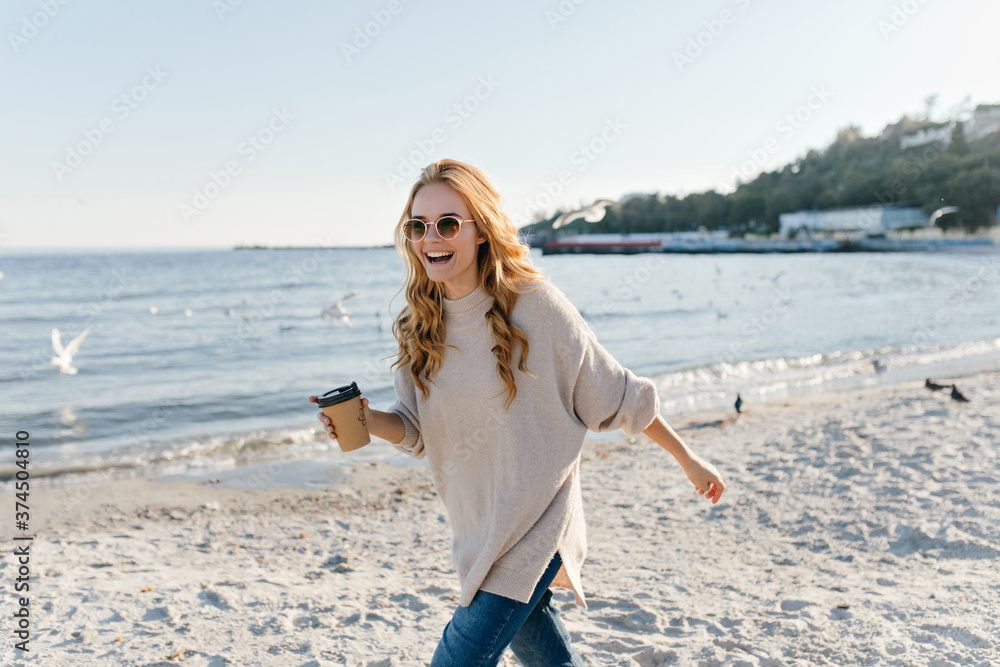 Active woman in sweater drinking tea at ocean coast. Outdoor portrait of joyful girl having fun at beach in autumn Active woman in sweater drinking tea at ocean coast. Outdoor portrait of joyful girl having fun at beach in autumn