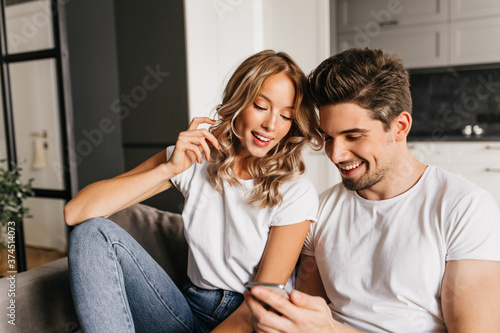 Fototapeta Naklejka Na Ścianę i Meble -  Happy couple with smart phone looking on the screen and smiling. Home photo of two young people enjoying day together and reading good news.