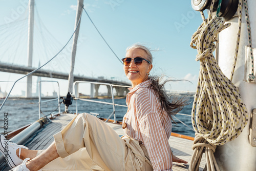 Happy mature woman wearing sunglasses on a sailboat and looking at camera. Senior female sitting on yacht deck and enjoying the voyage.