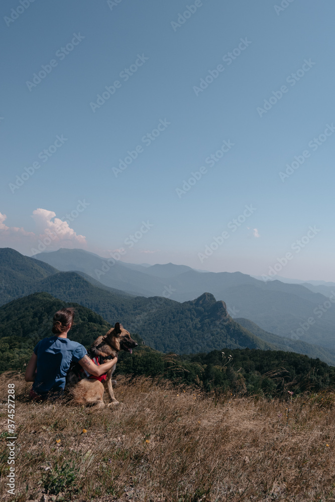 Fototapeta premium Dog traveler goes to the mountains with his best friend man and enjoys nature and beautiful scenery. A young traveller girl is sitting on top of a mountain with her German shepherd.