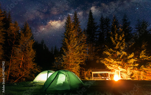  Stunning nature scenery. Two green tents under the amazing blue starry sky on the forest glade with fire. Travel recreation, outdoor activity, freedom concept background. Amazing wild nature