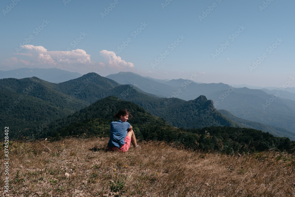 Naklejka premium Beautiful mountain and hilly landscape in warm, Sunny weather and the young tourist. A female traveler sits on top of a hill and looks at the surrounding mountains.